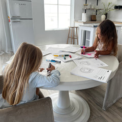 Two children sitting at a white table in a modern kitchen, coloring worksheets with markers.