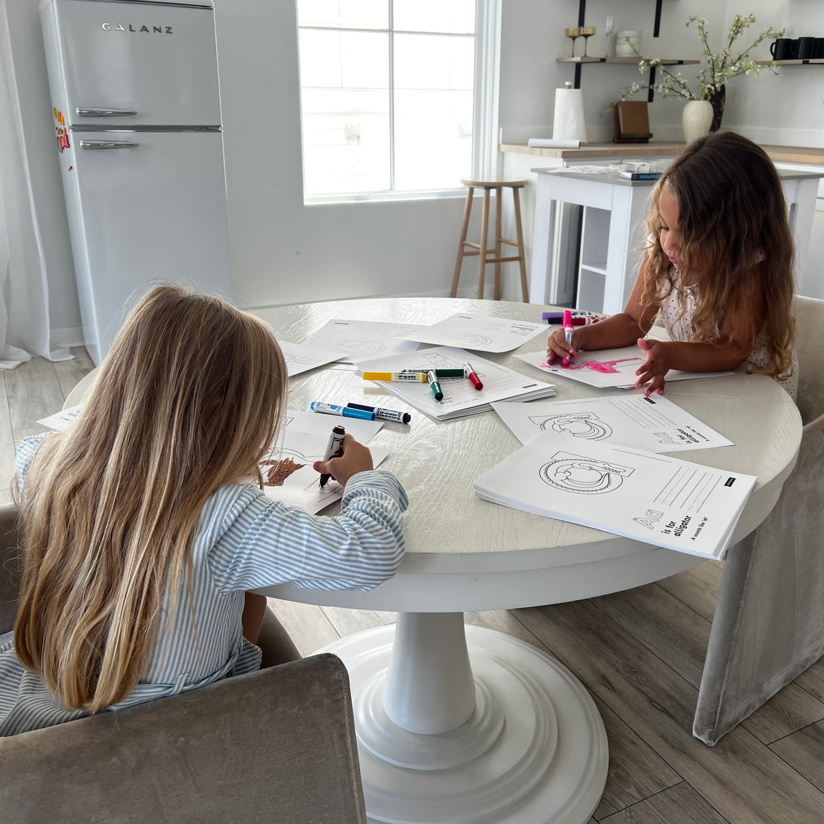 Two children sitting at a white table in a modern kitchen, coloring worksheets with markers.