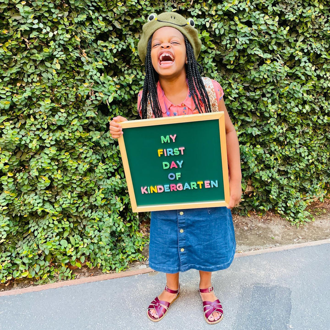 Child holding a sign saying 'My First Day of Kindergarten' in front of a green hedge.