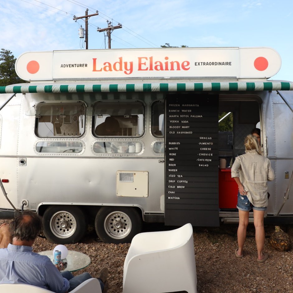 A vintage-style food truck with a white body, green-and-white awning, and large red 'Lady Elaine' signage, set outdoors on a dirt lot with people standing and sitting nearby.