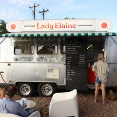 A vintage-style food truck with a white body, green-and-white awning, and large red 'Lady Elaine' signage, set outdoors on a dirt lot with people standing and sitting nearby.