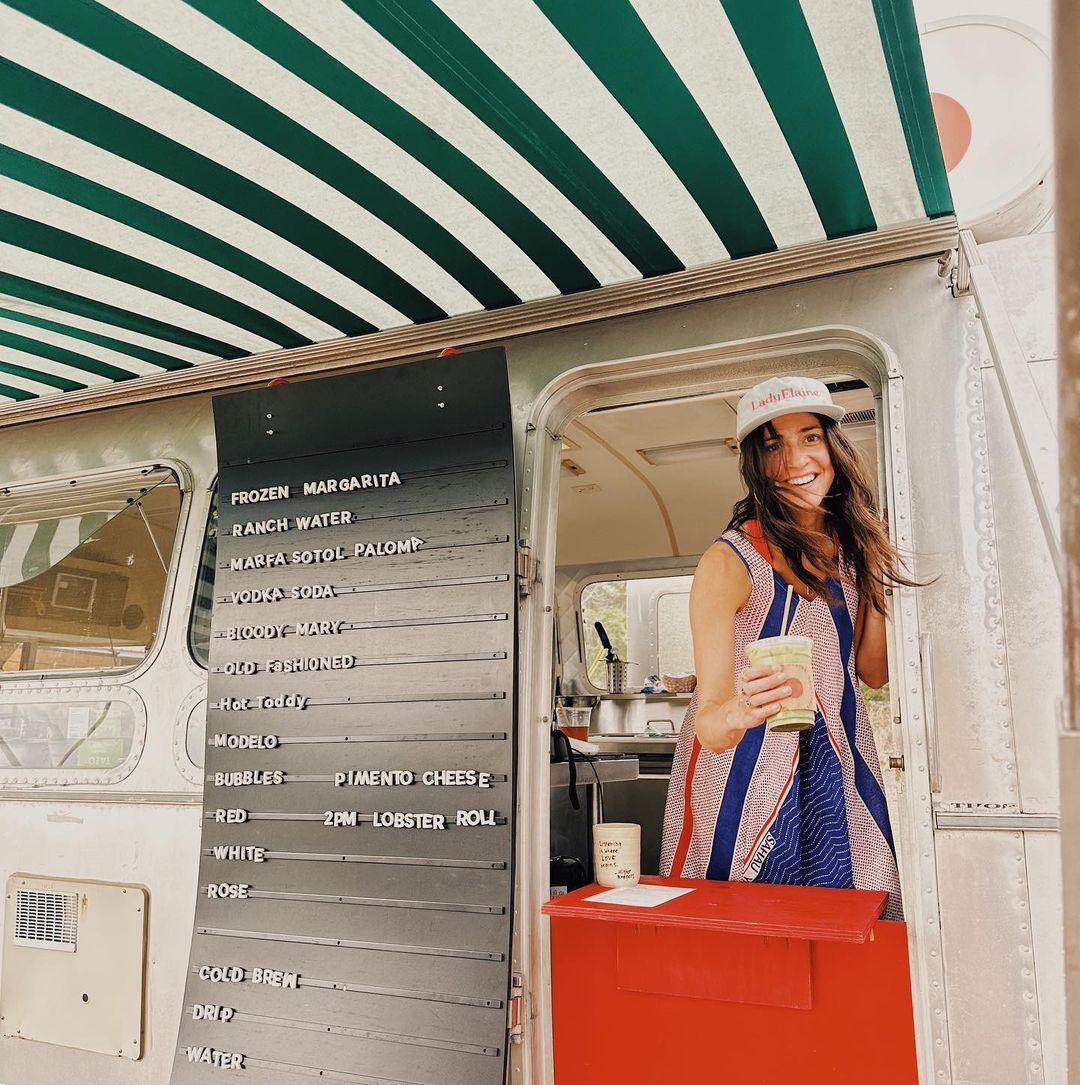 A woman stands in the doorway of a mobile food truck wearing a white cap with red lettering, a blue and white striped top, and holding a paper cup. The truck features a green and white striped awning, a silver exterior, and a black magnetic menu board listing items like frozen margarita, ranch water, and chicken salad.