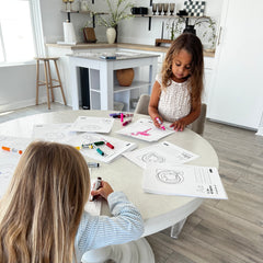 Two children sitting at a table with coloring worksheets and markers in a bright kitchen.