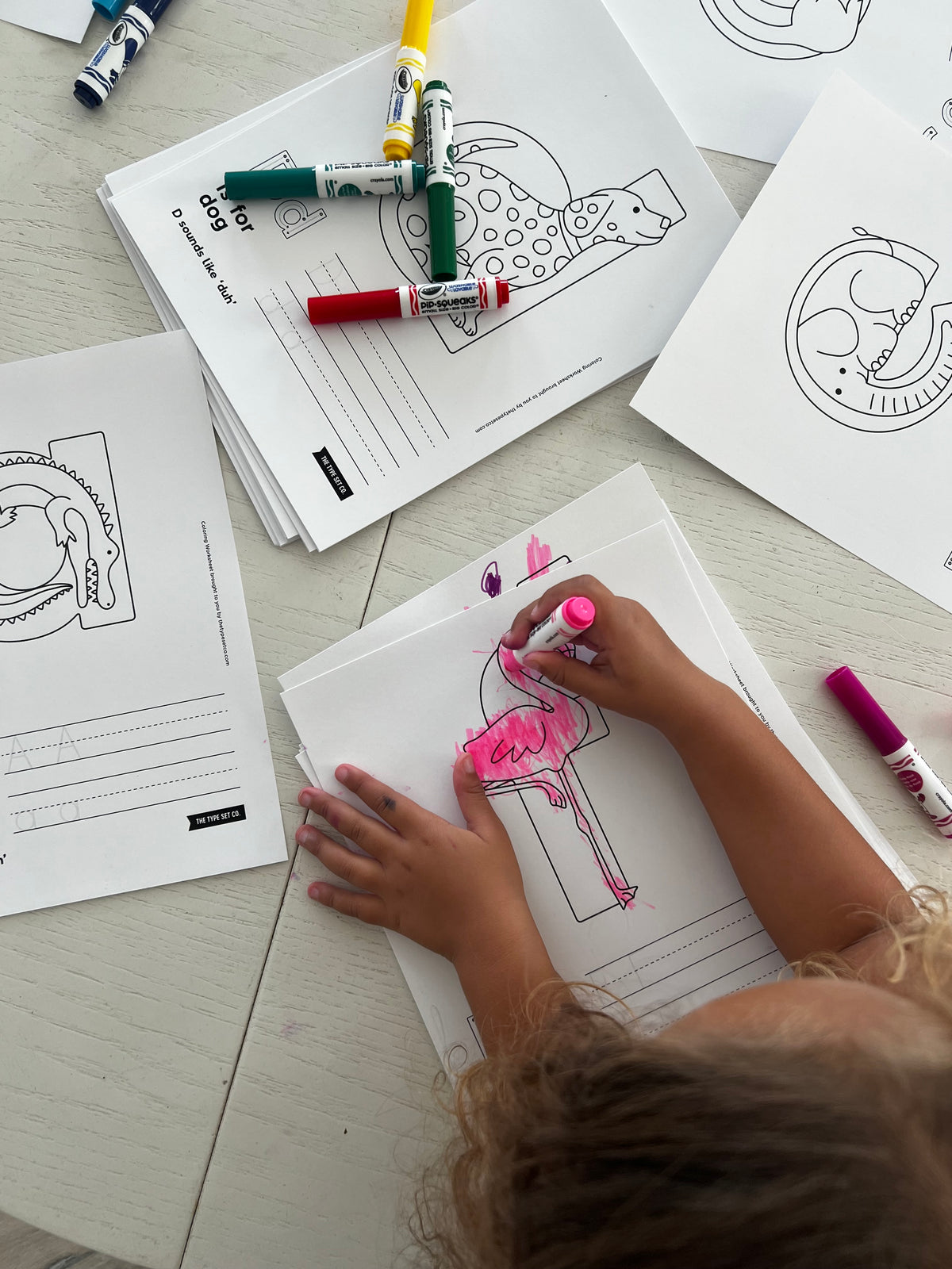 Child coloring a picture of a flamingo with markers on a table with other coloring alphabet worksheets scattered around
