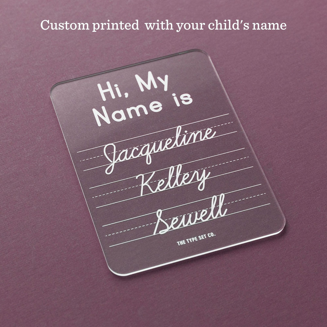 A clear acrylic name sign with rounded corners, printed with the text 'Hi, My Name is Jacqueline Kelly Sewell' in white. The sign is displayed on a textured dark purple background with a text overlay at the top reading 'Custom printed with your child's name'.