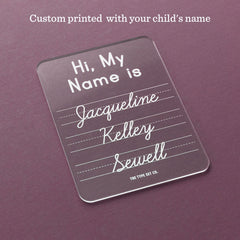 A clear acrylic name sign with rounded corners, printed with the text 'Hi, My Name is Jacqueline Kelly Sewell' in white. The sign is displayed on a textured dark purple background with a text overlay at the top reading 'Custom printed with your child's name'.