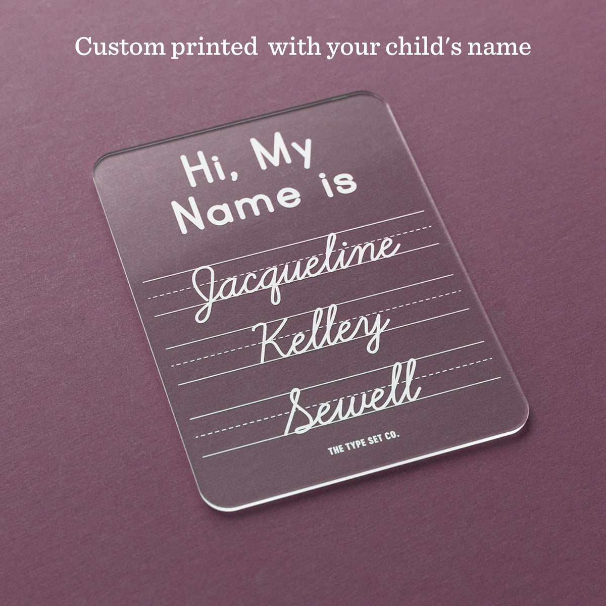 A clear acrylic name sign with rounded corners, printed with the text 'Hi, My Name is Jacqueline Kelly Sewell' in white. The sign is displayed on a textured dark purple background with a text overlay at the top reading 'Custom printed with your child's name'.