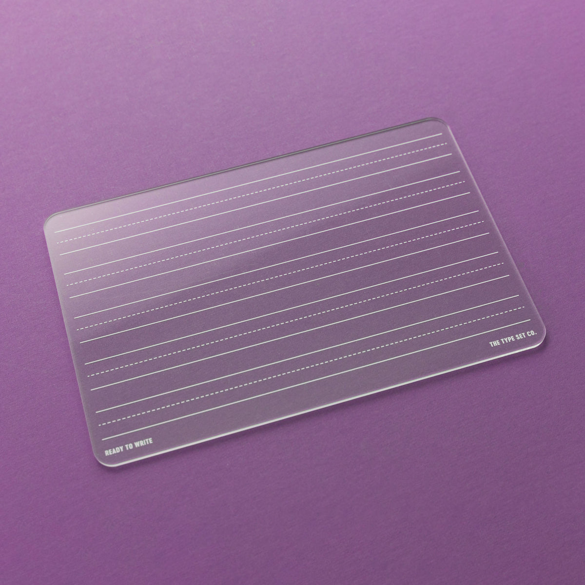 A clear rectangular acrylic board with rounded corners, engraved horizontal guide lines, and small printed text reading 'READY TO WRITE' and 'THE TYPE SET CO.' on a solid purple background.