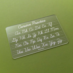 A clear acrylic cursive practice card with white lettering, shown at an angle on a textured olive green background.