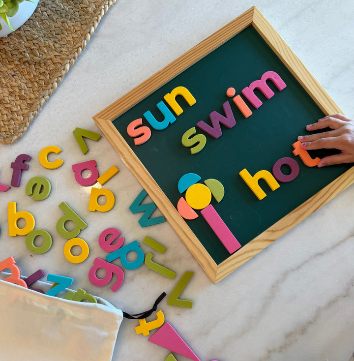 A wooden-framed magnetic letter board on a white marble surface, with colorful alphabet magnets spelling words like 'sun', 'swim', and 'hot'. A hand is placing a letter, and additional scattered letters and a fabric pouch are visible around it.