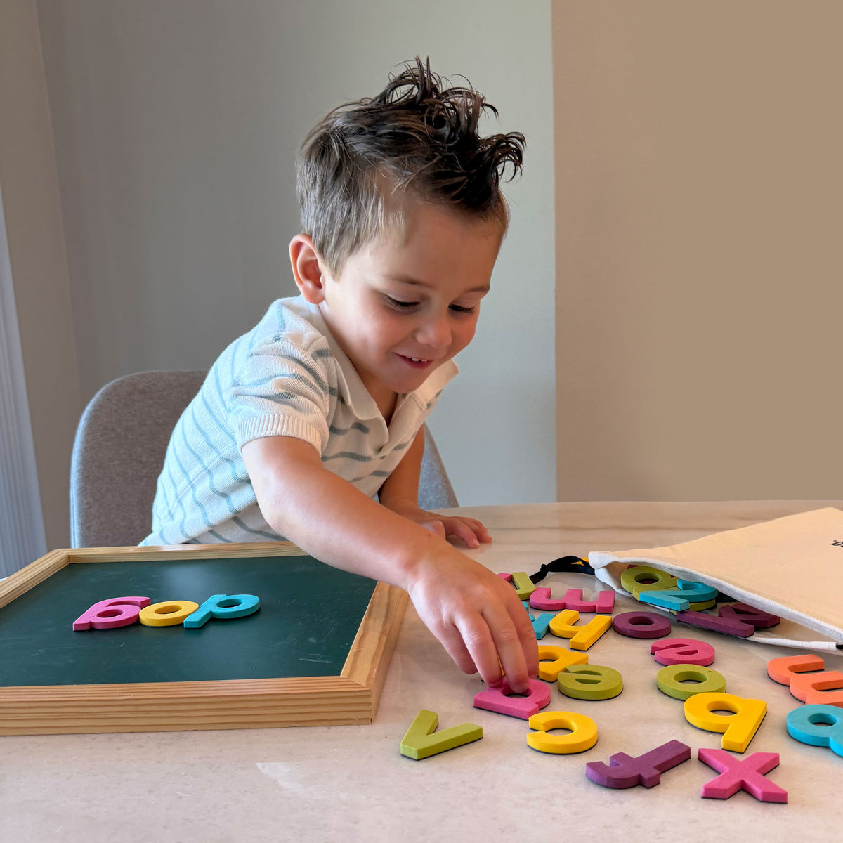 A young child sitting at a table, playing with colorful magnetic alphabet and number letters on a light-colored surface next to a small wooden-framed magnetic board.