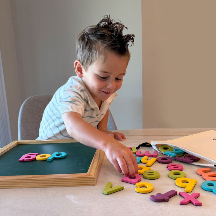 A young child sitting at a table, playing with colorful magnetic alphabet and number letters on a light-colored surface next to a small wooden-framed magnetic board.