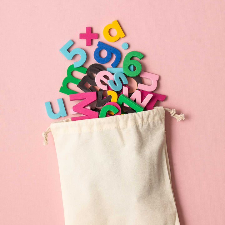 Rainbow magnetic letters spilling out of a canvas drawstring bag on a pink background