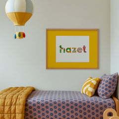 Framed artwork with a yellow mat featuring colorful animal-shaped letters spelling 'hazel' above a child's bed with patterned bedding and pillows.
