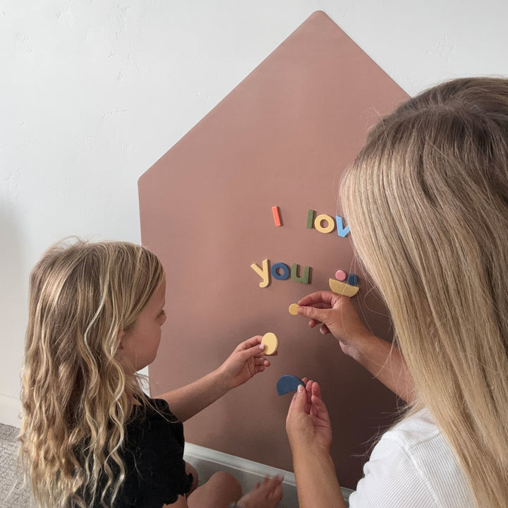 A woman and a young girl interact with colorful magnetic letter tiles spelling 'I love you' on a large tan house-shaped wall decal mounted on a white wall.