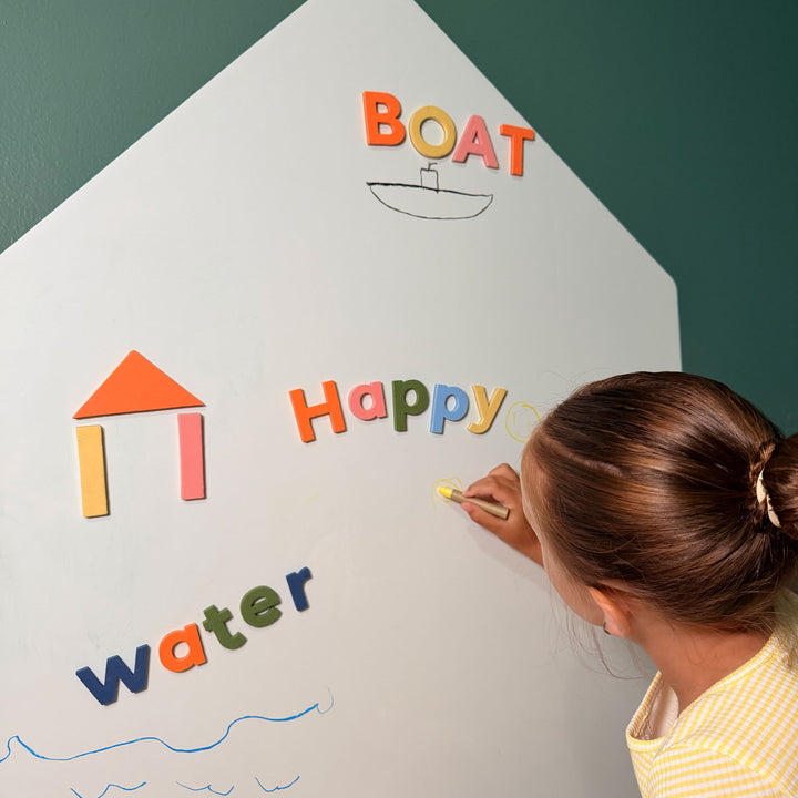 A child writes on a large white house-shaped board with colorful magnetic letters spelling words like "BOAT," "Happy," and "water," against a dark green wall.