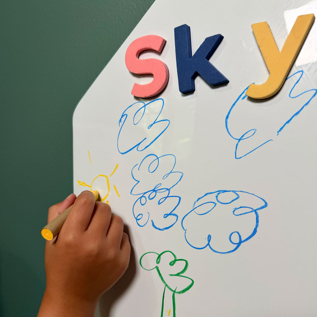 A child's hand holding a yellow wet-erase crayon draws on a white magnetic wall decal with colorful magnetic letters spelling 'sky' and blue cloud shapes against a dark green wall 