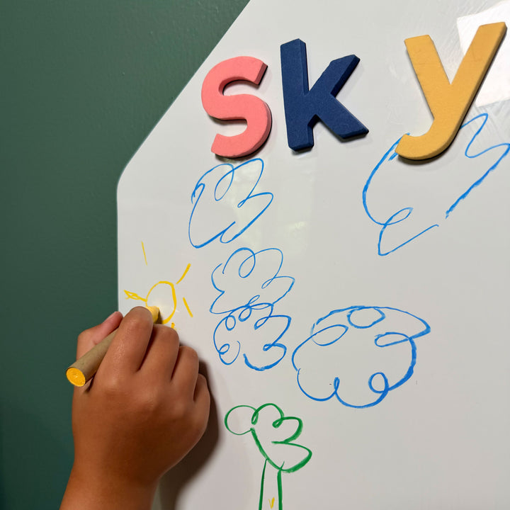 A child's hand holding a yellow wet-erase crayon draws on a white magnetic wall decal with colorful magnetic letters spelling 'sky' and blue cloud shapes against a dark green wall 