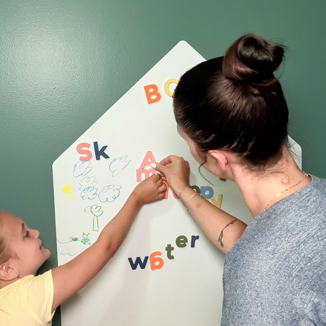 A woman and a young child are writing on a white house-shaped magnetic dry-erase wall decal with colorful letters against a green wall.