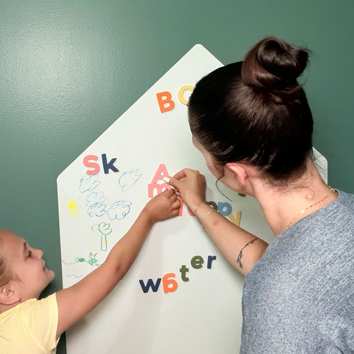 A woman and a young child are writing on a white house-shaped magnetic dry-erase wall decal with colorful letters against a green wall.