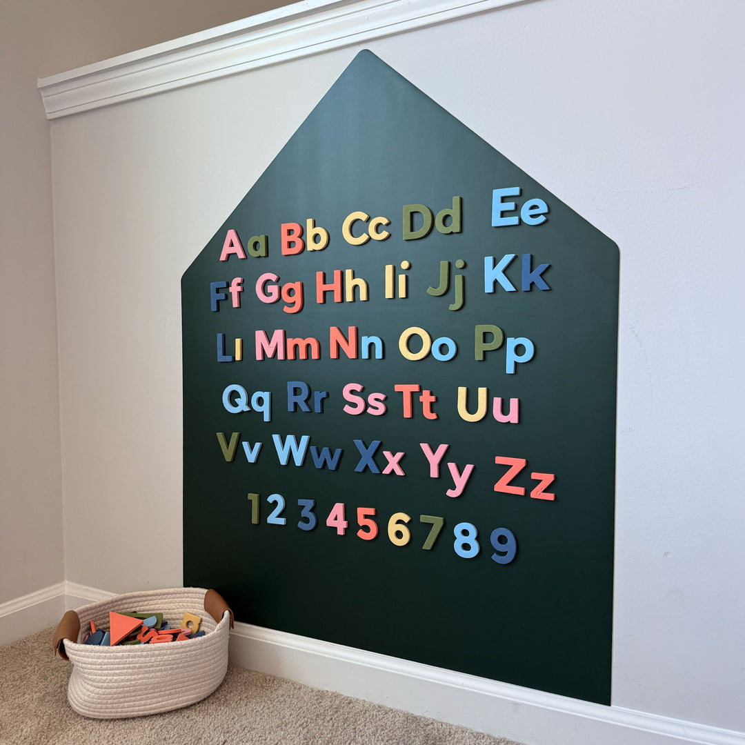 A house-shaped green magnetic wall decal with colorful magnetic letters and numbers arranged on the front. A woven basket of toys sits on the carpeted floor below.