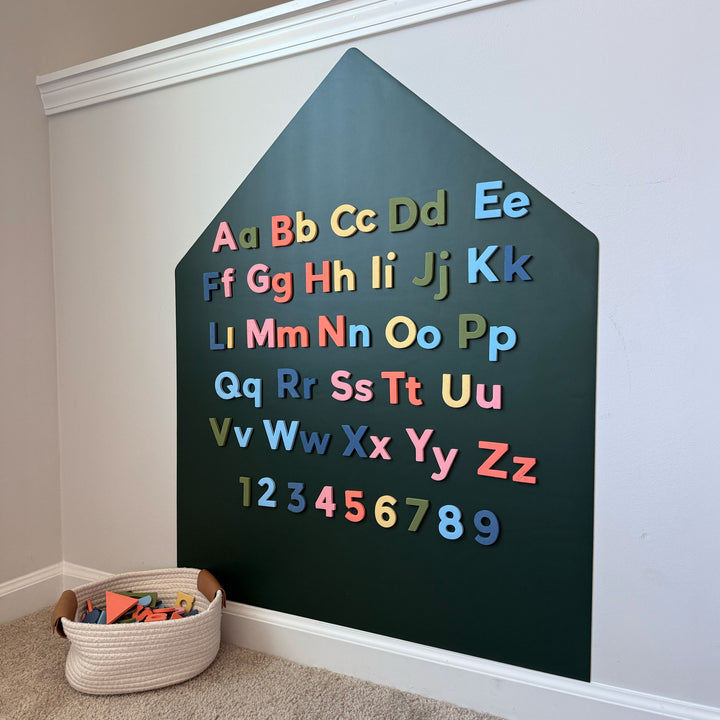 A house-shaped green magnetic wall decal with colorful magnetic letters and numbers arranged on the front. A woven basket of toys sits on the carpeted floor below.