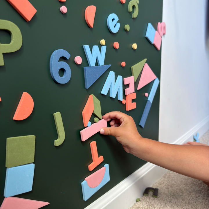 A child's hand placing a pink foam magnetic shape on a dark green magnetic wall decal covered with colorful foam letters, numbers, and shapes.