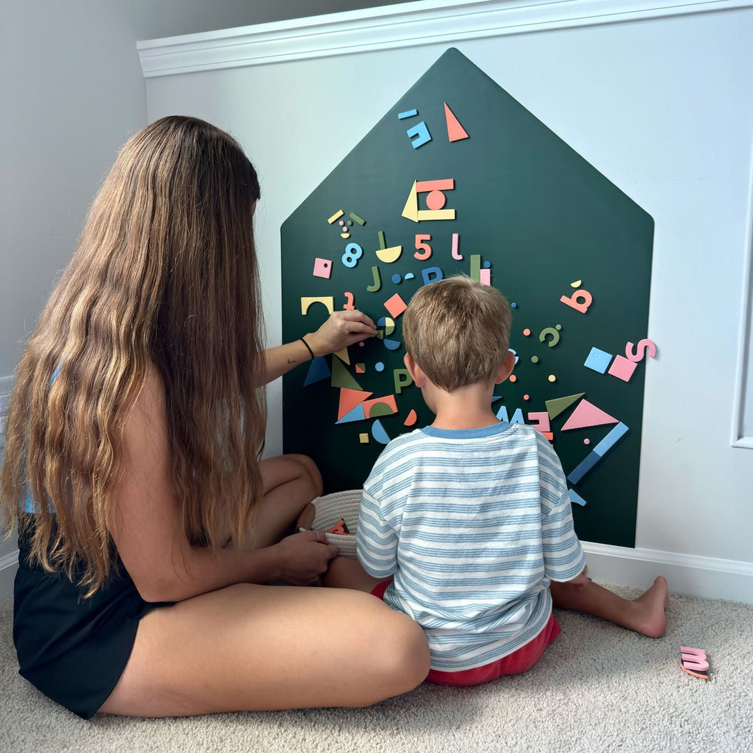 A woman and a young boy sit on a carpeted floor in front of a house-shaped magnetic wall decal, playing with colorful magnetic letters, numbers, and shapes.