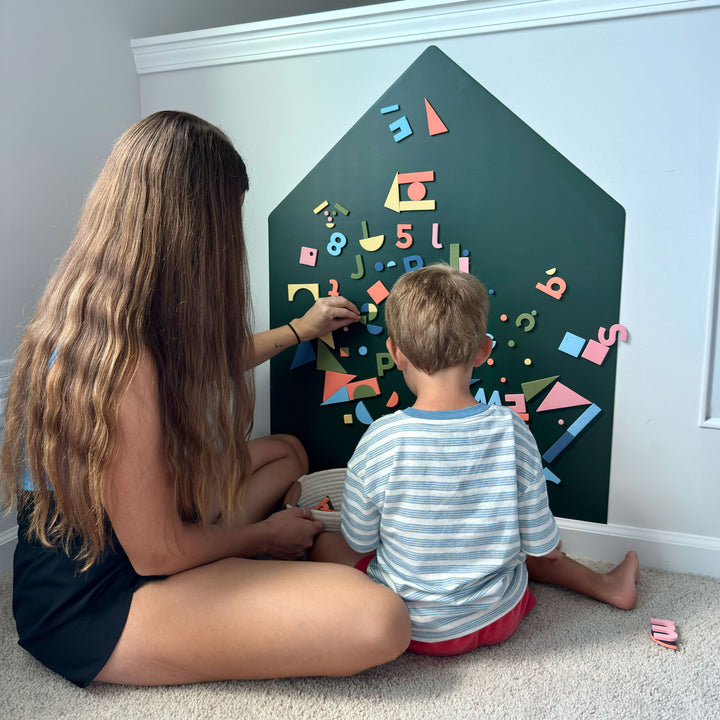 A woman and a young boy sit on a carpeted floor in front of a house-shaped magnetic wall decal, playing with colorful magnetic letters, numbers, and shapes.