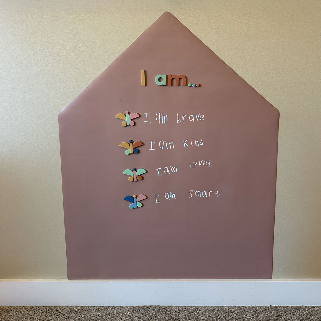 A house-shaped chalkboard wall decal piece mounted on a beige wall above a white baseboard. The board is a pinky-tan color and features colorful magnetic letters and magnetic shapes with words of affirmation.