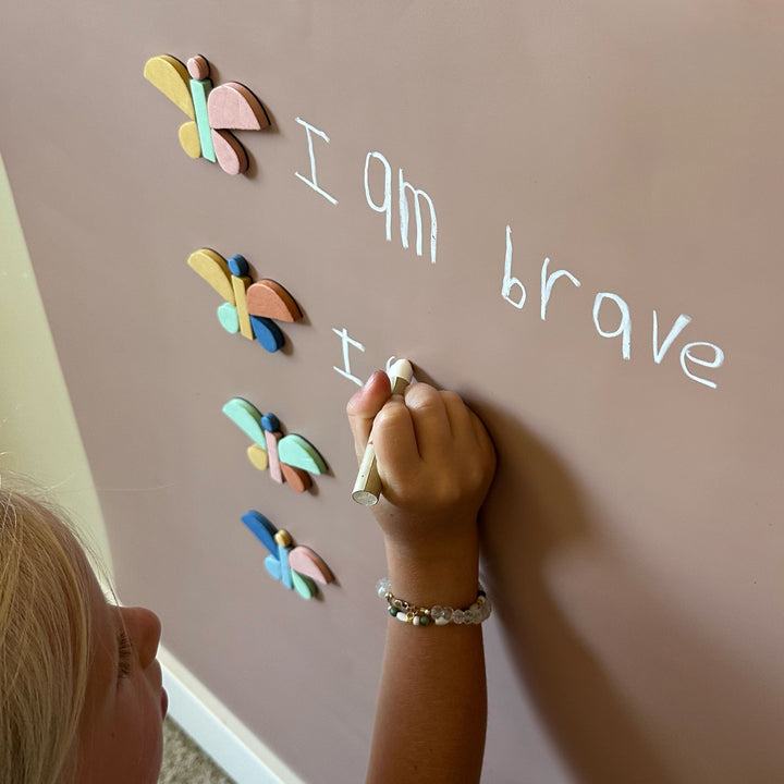 A child's hand writes on a tan magnetic wall decal next to colorful butterfly magnets and the words 'I am brave' in white marker.